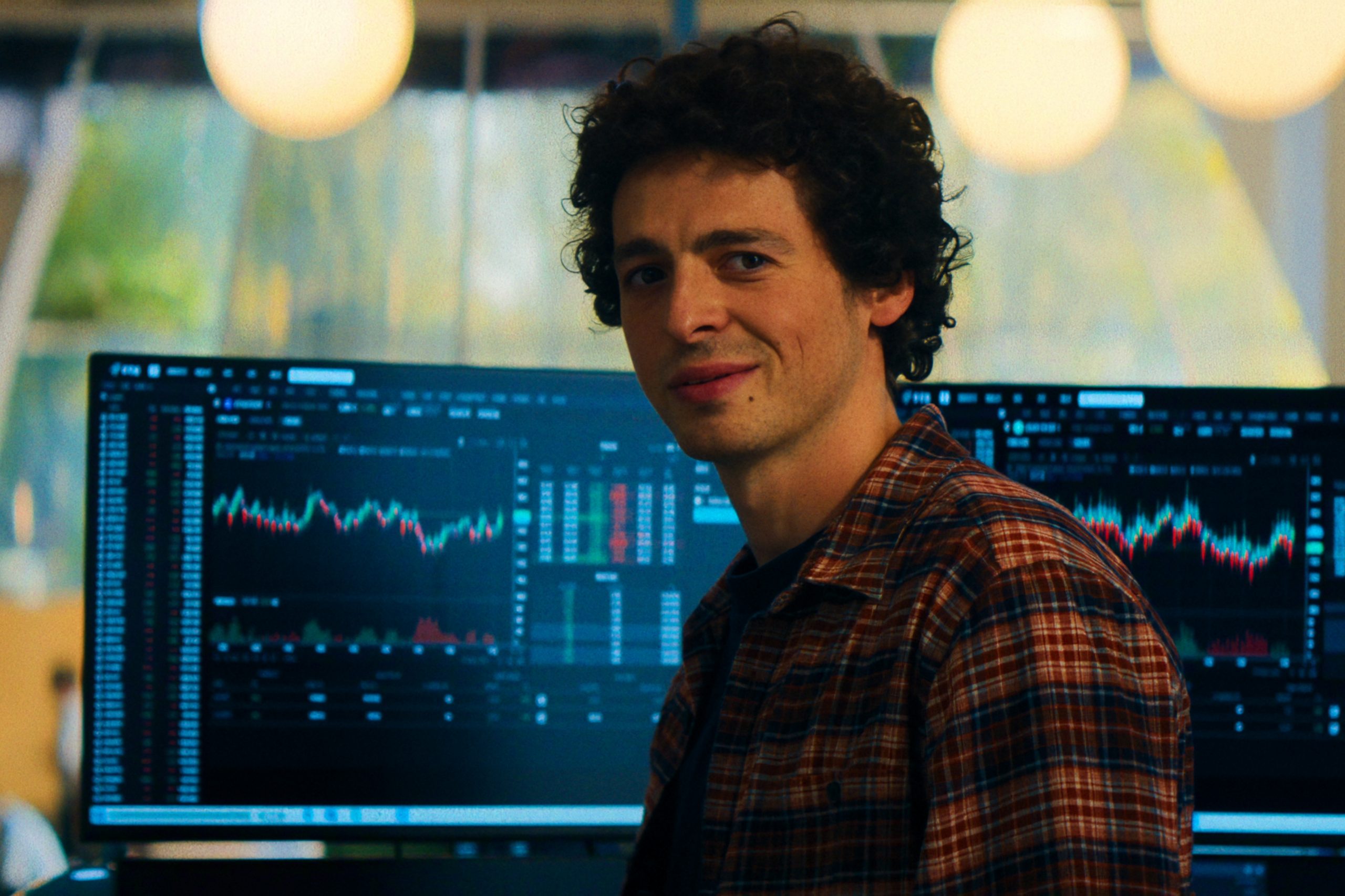 "A man in a plaid shirt sits in front of computer monitors displaying financial charts and stock data in a modern office with large windows and warm lighting."
